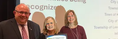 people standing on a stage holding an award certificate, with a large presentation screen behind them displaying the message “Congratulations to the Age-Friendly Communities Being Recognized” along with a list of municipalities.