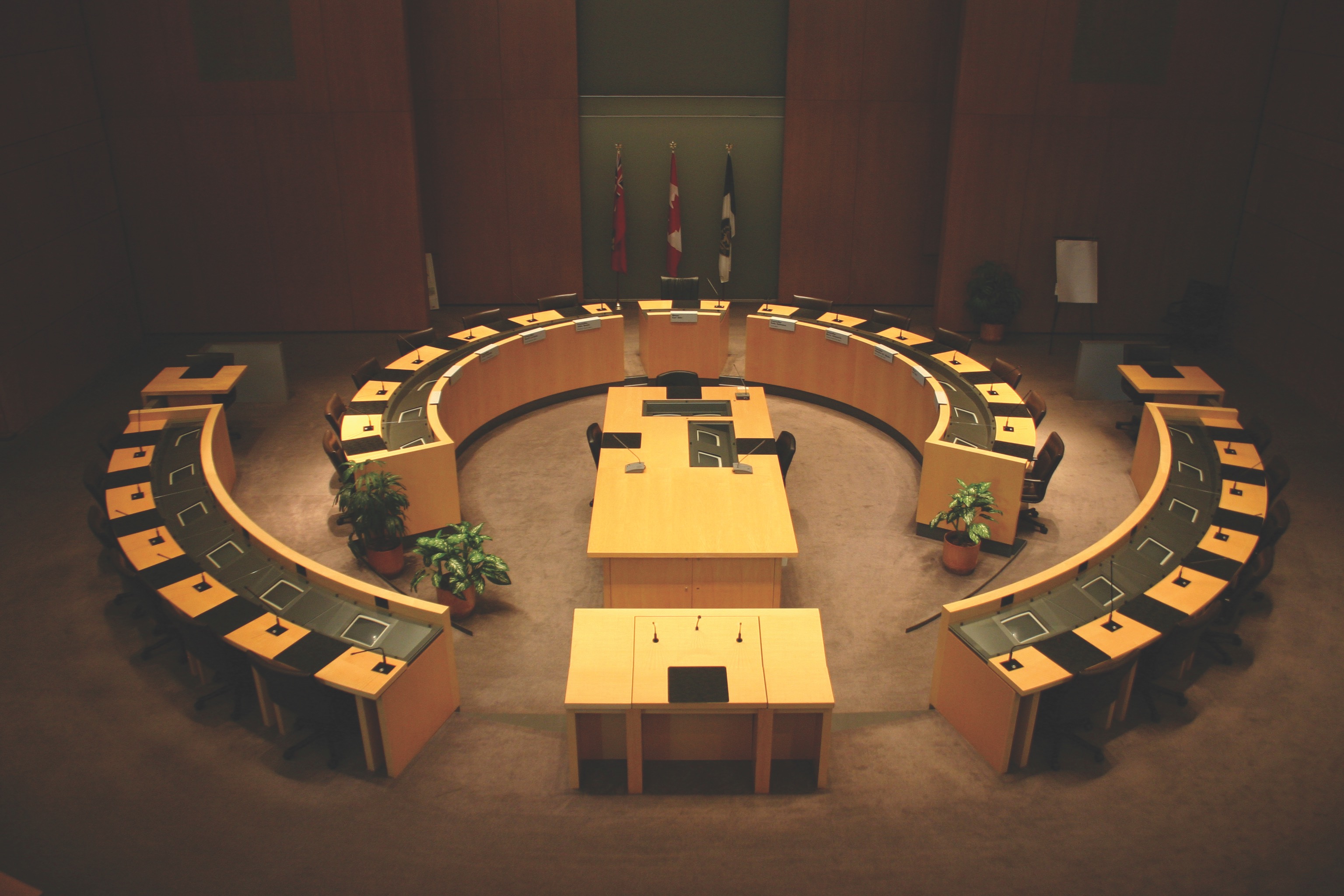 Council chamber with two semi-circular rows of desks facing a central podium and flags on the back wall.