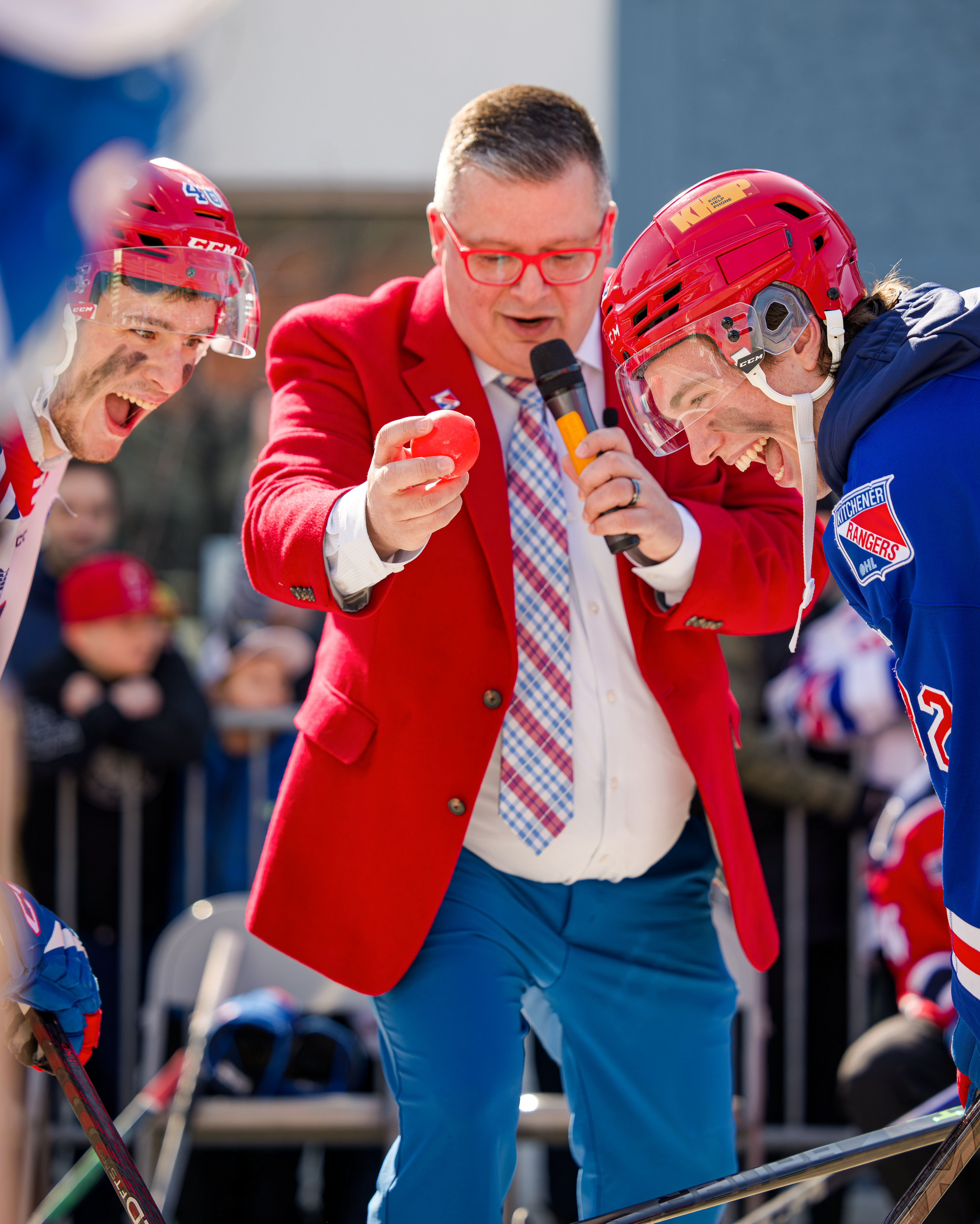 Two Kitchener Rangers hockey players face off on either edge of the image while an announcer drops a ball in the centre