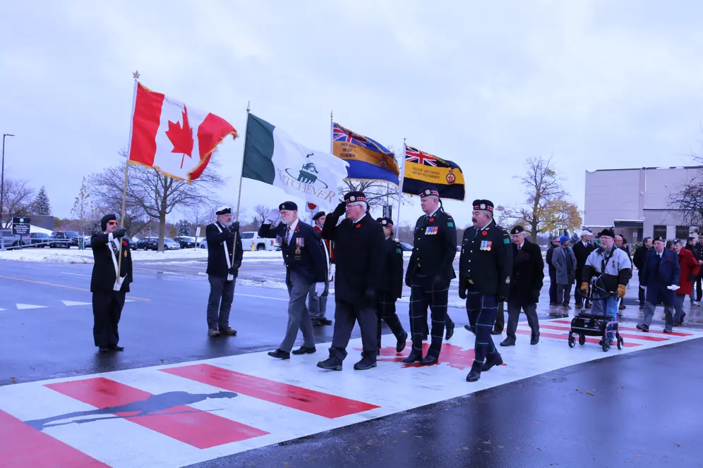 Veterans and community members march across a crosswalk with flags on a cloudy day.